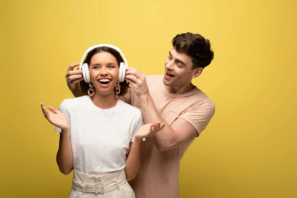 Homem bonito colocando fones de ouvido em menina alegre no fundo amarelo — Fotografia de Stock