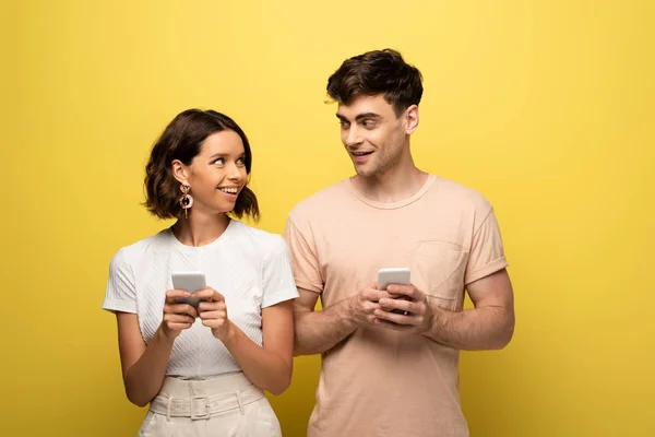 Hombre y mujer positivos que se miran entre sí mientras usan teléfonos inteligentes sobre fondo amarillo - foto de stock