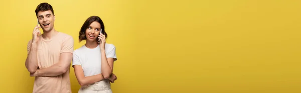 Panoramic shot of young man and woman talking on smartphones on yellow background — Stock Photo