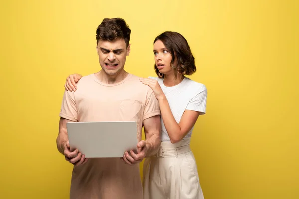 Pretty girl calming irritated boyfriend using laptop on yellow background — Stock Photo