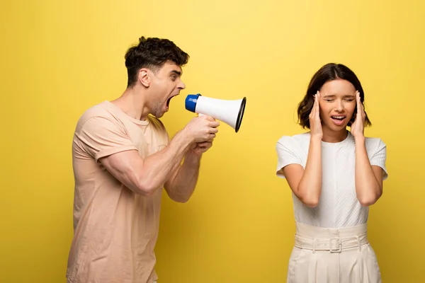 Wütender junger Mann streitet im Lautsprecher über Freundin, die Ohren mit Händen auf gelbem Hintergrund bedeckt — Stockfoto
