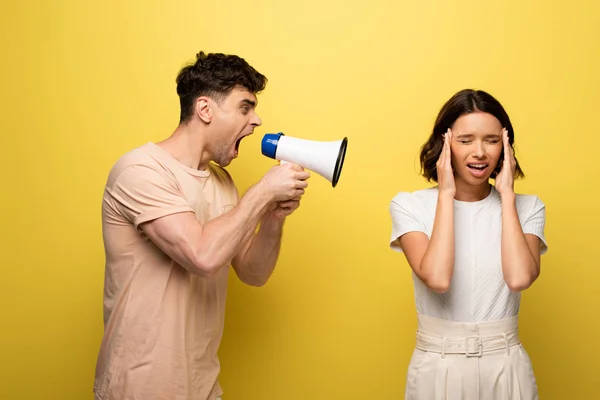 Angry young man quarreling in loudspeaker at girfriend covering ears with hands on yellow background — Stock Photo