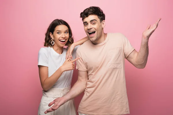 Cheerful young man showing shrug gesture while standing near pretty girlfriend on pink background — Stock Photo