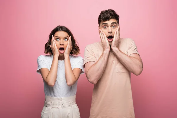 Homem chocado e mulher de mãos dadas perto do rosto enquanto olha para a câmera no fundo rosa — Fotografia de Stock