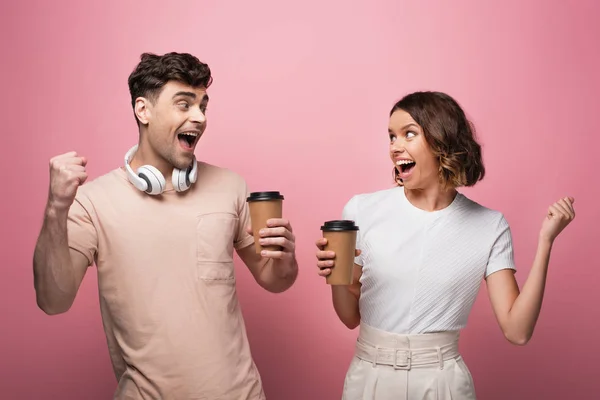 Happy man and woman showing yes gestures while holding paper cups and looking at each other on pink background — Stock Photo