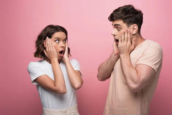 Schockierter Mann und Frau halten sich die Hände in der Nähe von Gesichtern, während sie sich auf rosa Hintergrund anschauen — Stockfoto