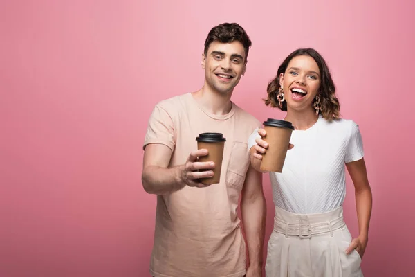 Happy man and woman hoding paper cups and smiling at camera on pink background — Stock Photo