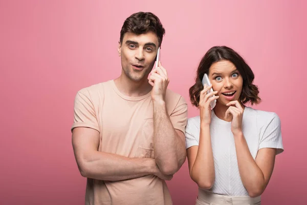 Homme et femme joyeux parlant sur les smartphones tout en regardant la caméra sur fond rose — Photo de stock