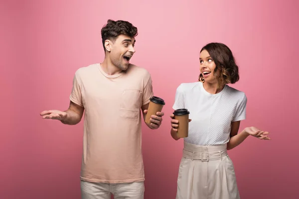 Sorridente homem e mulher mascando encolher de ombros gestos enquanto olhando um para o outro no fundo rosa — Fotografia de Stock