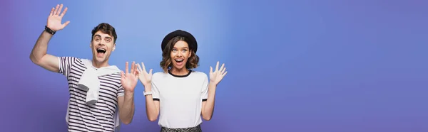 Panoramic shot of excited man and woman waving hand and smiling at camera on blue background — Stock Photo