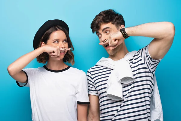 Fröhlicher Mann und Frau, die sich anschauen, während sie die Finger mit gezogenen Schnurrbärten vor blauem Hintergrund halten — Stockfoto