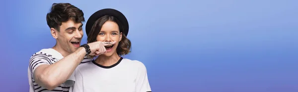 Plano panorámico de hombre alegre sosteniendo el dedo con bigote dibujado cerca de la cara de la joven sobre fondo azul - foto de stock