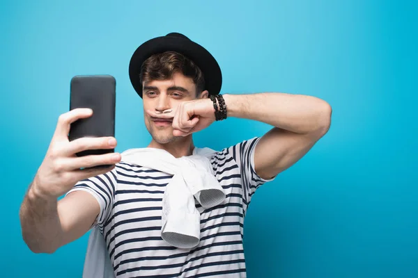 Hombre alegre tomando selfie con teléfono inteligente mientras sostiene el dedo con bigote dibujado cerca de la cara sobre fondo azul - foto de stock