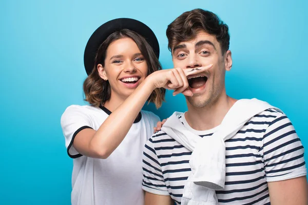 Chica alegre sosteniendo el dedo con bigote dibujado cerca de la cara del hombre joven sobre fondo azul - foto de stock