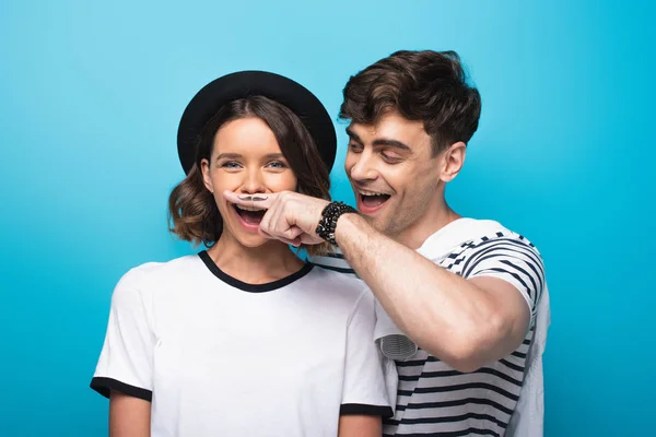 Cheerful man holding finger with drawn mustache near face of young woman on blue background — Stock Photo