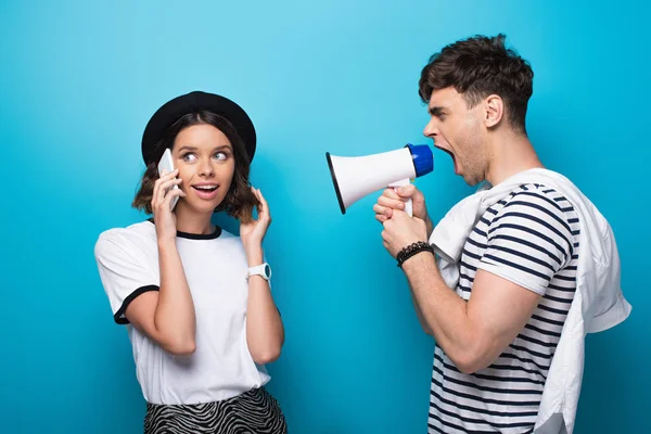 Irritated man quarreling in loudspeaker at girlfriend talking on smartphone on blue background — Stock Photo