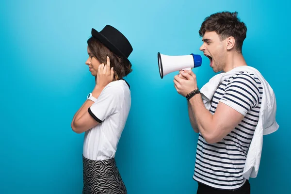 Angry man quarreling in loudspeaker at girlfriend plugging ears with fingers on blue background — Stock Photo