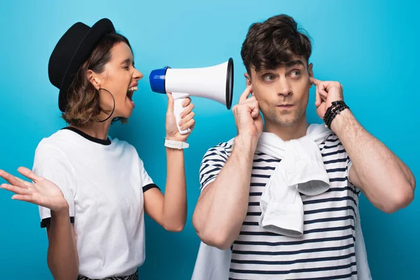 Chica enojada peleando en altavoz en novio tapando las orejas con los dedos sobre fondo azul - foto de stock