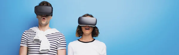 Панорамный снимок молодого человека и женщины с помощью наушников виртуальной реальности на синем фоне — стоковое фото