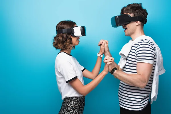 Joven hombre y mujer tomados de la mano mientras usa auriculares de realidad virtual sobre fondo azul - foto de stock