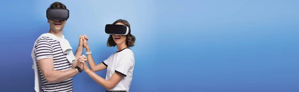 Panoramic shot of young man and woman holding hands while using virtual reality headsets on blue background — Stock Photo