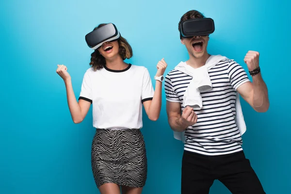 Alegre hombre y mujer bailando mientras usa auriculares de realidad virtual sobre fondo azul - foto de stock