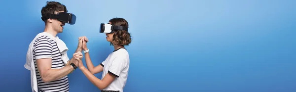 Panoramic shot of young man and woman holding hands while using virtual reality headsets on blue background — Stock Photo