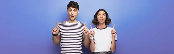 Panoramic shot of cheerful man and woman looking up and pointing with fingers on blue background — Stock Photo