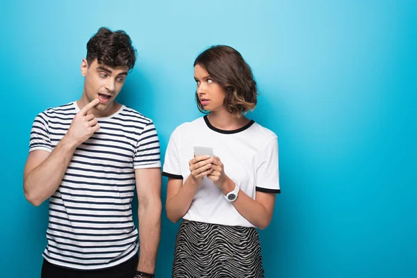 Thoughtful young man looking at smartphone in hands of pretty girl on blue background — Stock Photo