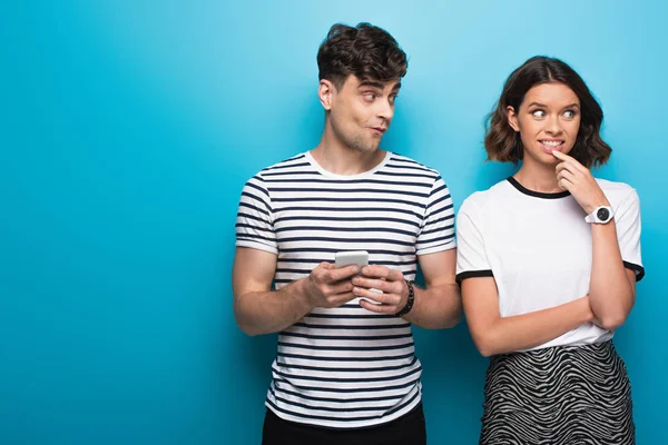 Young man holding smartphone and looking at thoughtful girl on blue background — Stock Photo