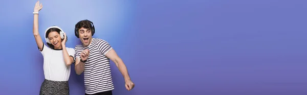 Панорамний знімок веселого чоловіка і жінки, що танцює під час прослуховування музики в навушниках на синьому фоні — стокове фото
