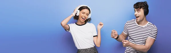 Panoramic shot of excited man and woman dancing while listening music in headphones on blue background — Stock Photo