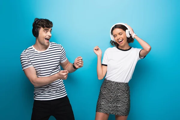 Alegre hombre y mujer bailando mientras escucha música en auriculares sobre fondo azul - foto de stock
