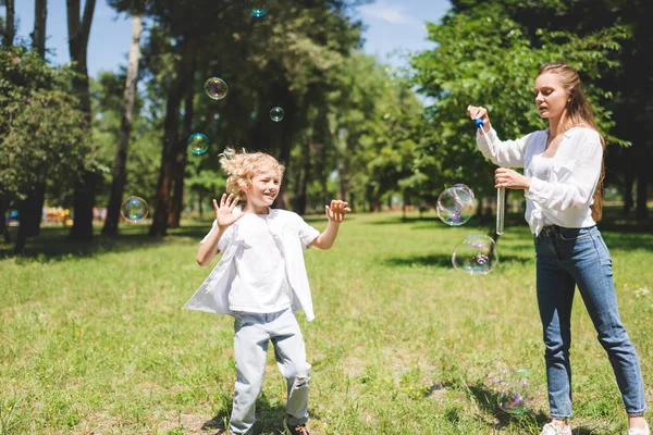 Красивая мама раздувает мыльные пузыри для очаровательного сына в парке — стоковое фото