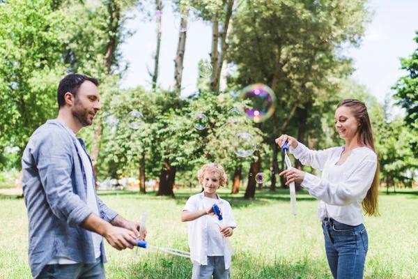 Mother, father and son in casual clothes playing with soap bubbles in park — Stock Photo