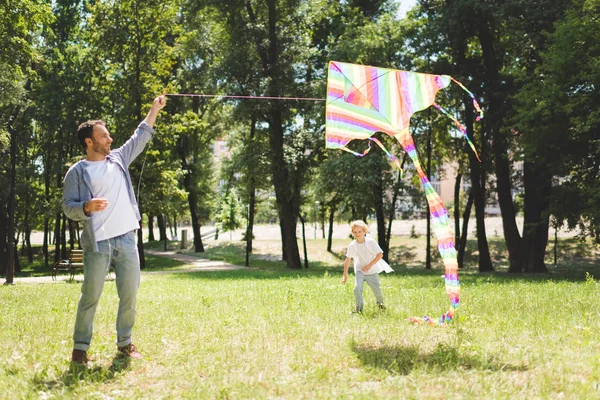 Padre y adorable hijo jugando con colofrul volando cometa en parque - foto de stock