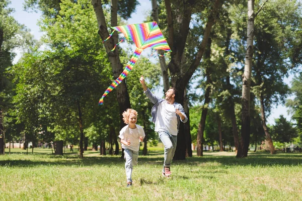 Счастливый отец и очаровательный сын играют с колофруктовым летучим змеем в парке — стоковое фото