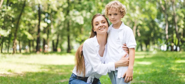 Panoramic shot of beautiful smiling mom hugging son in park during daytime — Stock Photo