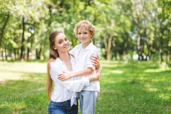 Hermosa feliz mamá abrazando hijo en el parque y mirando a la cámara - foto de stock