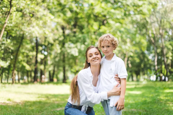 Schöne lächelnde Mutter umarmt Sohn im Park und schaut in die Kamera — Stockfoto