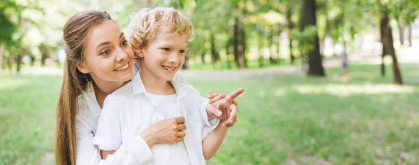 Tiro panorâmico de mãe bonita abraçando filho adorável apontando com o dedo no espaço cópia no parque — Fotografia de Stock
