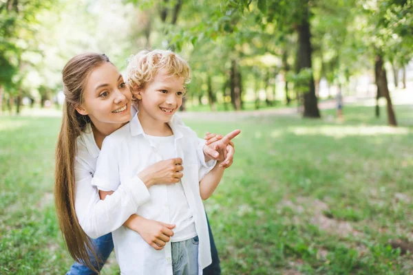 Bella madre abbracciare adorabile figlio che punta con il dito a copiare spazio nel parco — Foto stock