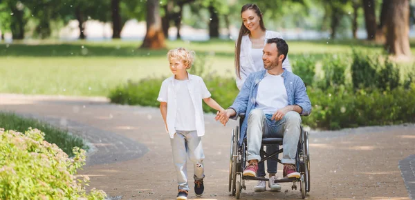 Panoramic shot of beautiful mother rolling wheelchair with disabled father in park near son — Stock Photo