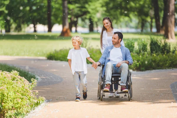 Madre rodando silla de ruedas con padre discapacitado en parque cerca de hijo - foto de stock