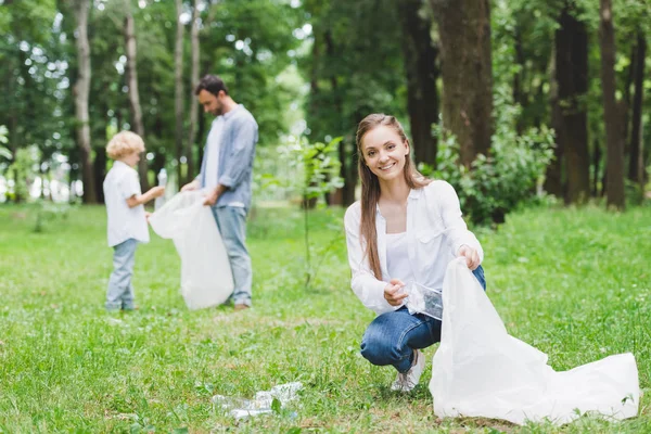 Smiling mother, father and son picking up plastic bottles in bags in park — Stock Photo
