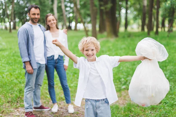 Feliz hijo sosteniendo bolsa de basura cerca de los padres en el parque - foto de stock