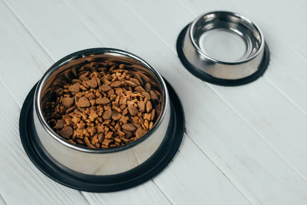 Cuenco de metal con comida para mascotas y cuenco vacío en superficie de madera blanca - foto de stock