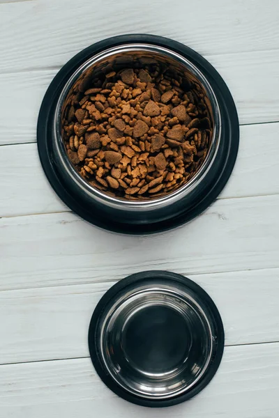 Vista superior de cuenco de metal con comida para mascotas y cuenco vacío en superficie de madera - foto de stock