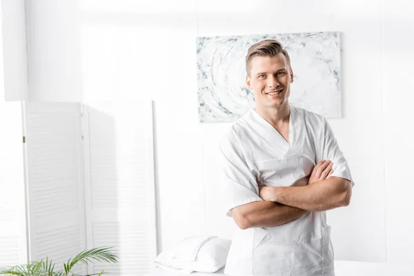 Smiling masseur standing with crossed arms and looking at camera in clinic — Stock Photo