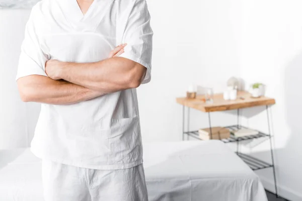 Vue recadrée du masseur debout avec les bras croisés et regardant la caméra à la clinique — Photo de stock